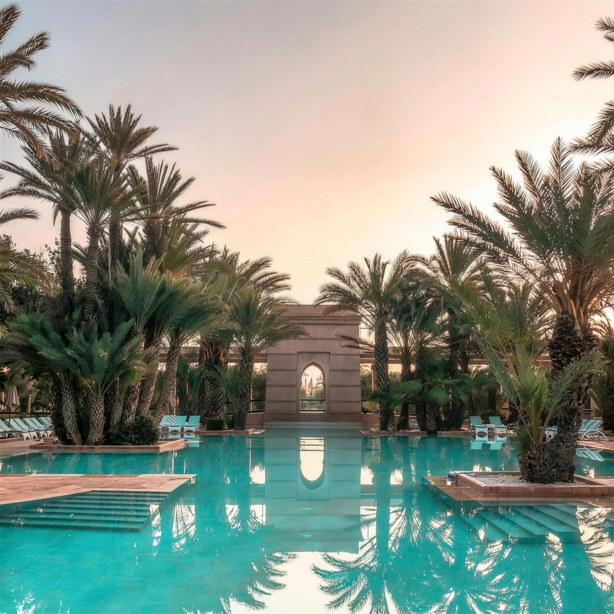 Resort pool lined with palm trees and an arched pavilion at sunset